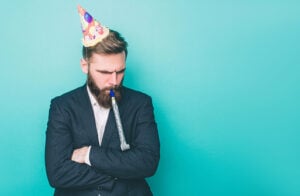 Sad guy is standing and looking down. He is upset. Man is holding a wistle in his mouth and has a birthday hat on the head. Isolated on blue background.