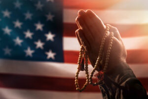 prayer Memorial day, veterans day. Male hands folded in prayer, holding a rosary. The American flag is in the background. The concept amerikanskih holidays and religion