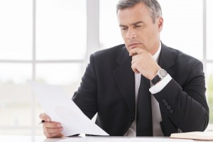 Businessman examining contract. Thoughtful mature man in formalwear holding hand on chin while examining document and sitting at working place