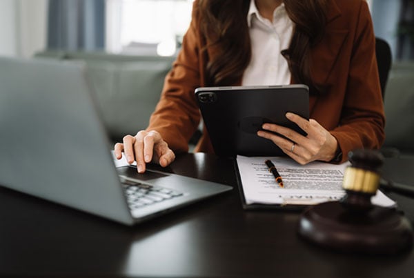A legal professional working on a paper document, a laptop, and a tablet; a gavel is on the desk